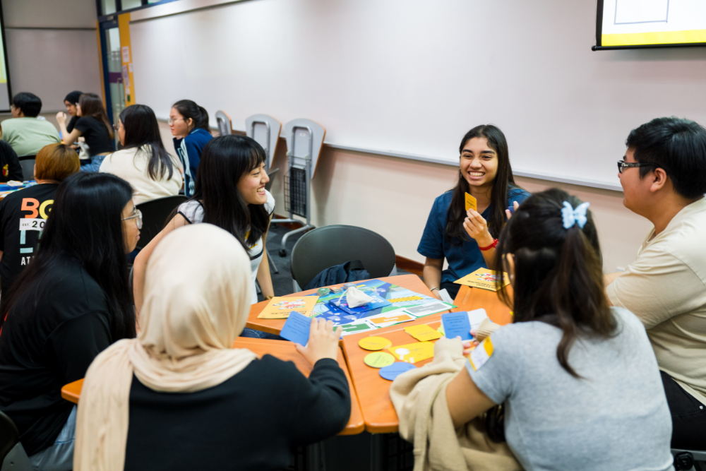 Students at a table play a card game using blue and yellow cards, along with a game board.